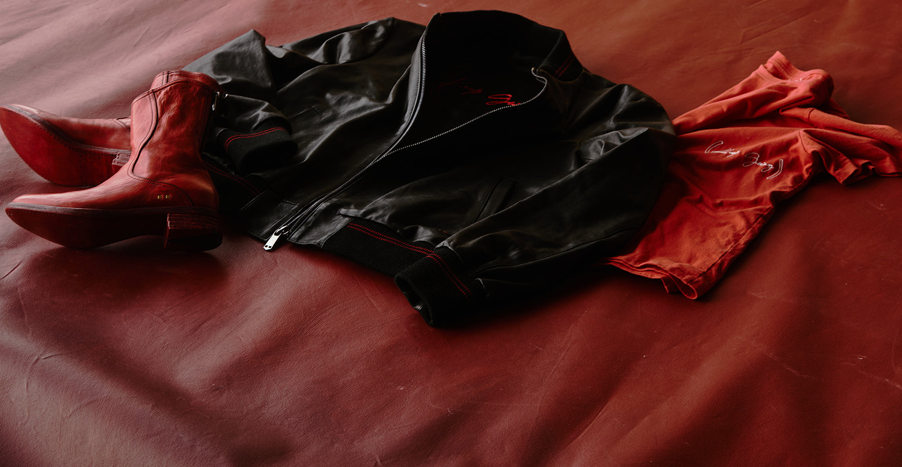 Red Savas Boots, a savas x Red bomber jacket and savas x red t shirt laid out on a brick red leather background.